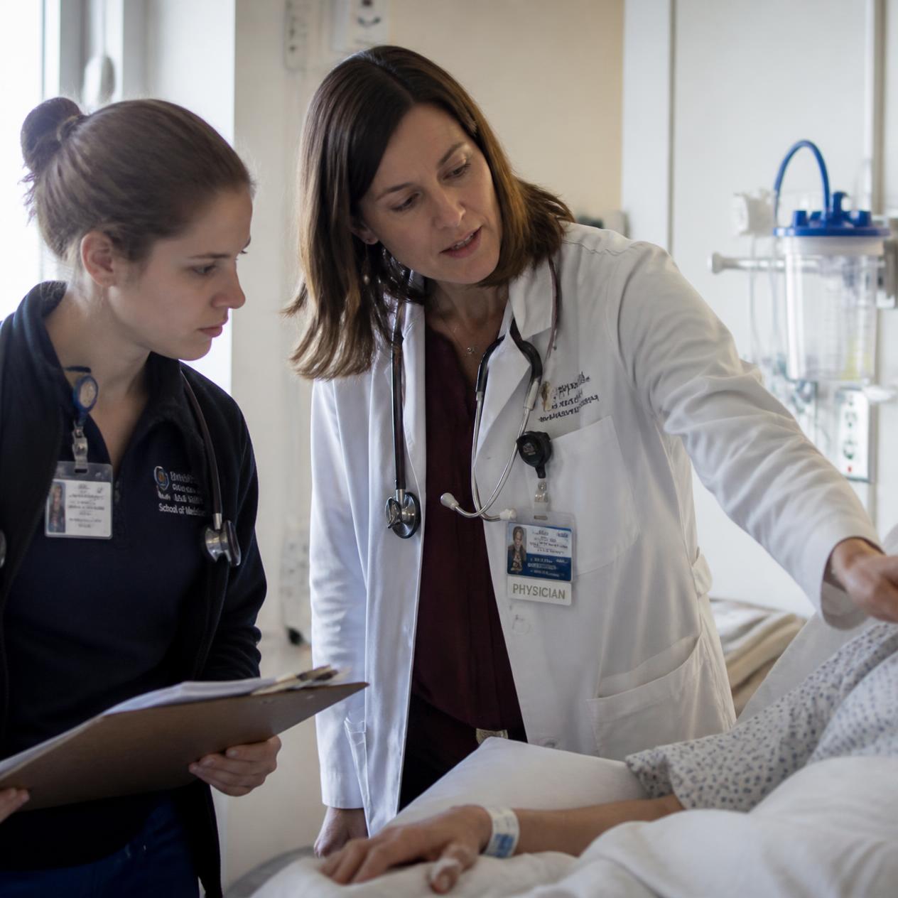 An attending physician teaches a medical student at a patient's bedside.